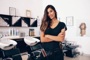 istockphoto-853924196-612×612 Female hairdresser in salon holding scissors in hand. Smiling young hairdresser standing in salon.